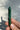 A polished green malachite crystal point held in a person's hand against a light gray textured background with a soft shadow.